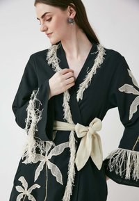 Svart rock med vit frans längs kanterna och intrikata blommönster i spets. Inkluderar ett beige tygbälte knutet i en rosett.