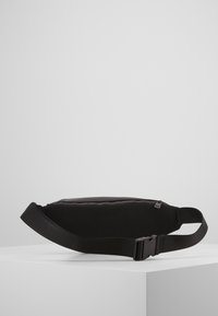 Czarna saszetka z gładkim przodem, teksturowanym siatkowym tyłem, regulowanym paskiem oraz zamkiem błyskawicznym zapewniającym bezpieczne przechowywanie.