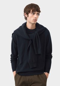 Maglione di lana navy con scollo a girocollo e un ampio colletto drappeggiato. Il modello lo abbina a pantaloni verde oliva, con le mani inserite nelle tasche.