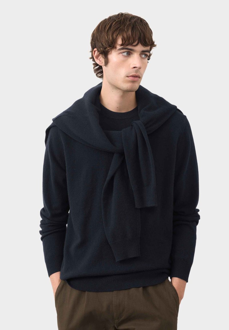 Maglione di lana navy con scollo a girocollo e un ampio colletto drappeggiato. Il modello lo abbina a pantaloni verde oliva, con le mani inserite nelle tasche.