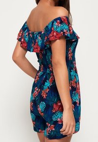Bloemenprint off-shoulder romper met een aansluitend lijfje, gerafelde randen en kleurrijke patronen van bloemen en tropische bladeren op een marineblauwe achtergrond.