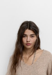 Beige strikket sweater med en dyb V-hals, der har en blød tekstur. Accentueret med en sølv kædehalssmykke. Ensfarvet baggrund.