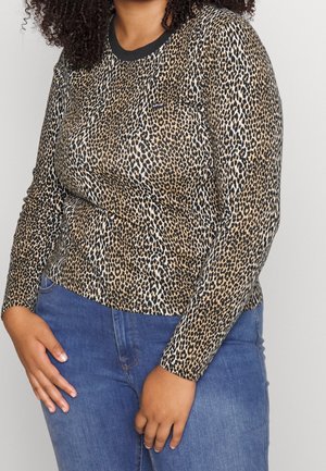 Vrouw in een lange mouwen luipaardprint top met zwarte kraag en blauwe jeans, met haar handen nabij haar heupen tegen een effen achtergrond geposeerd.