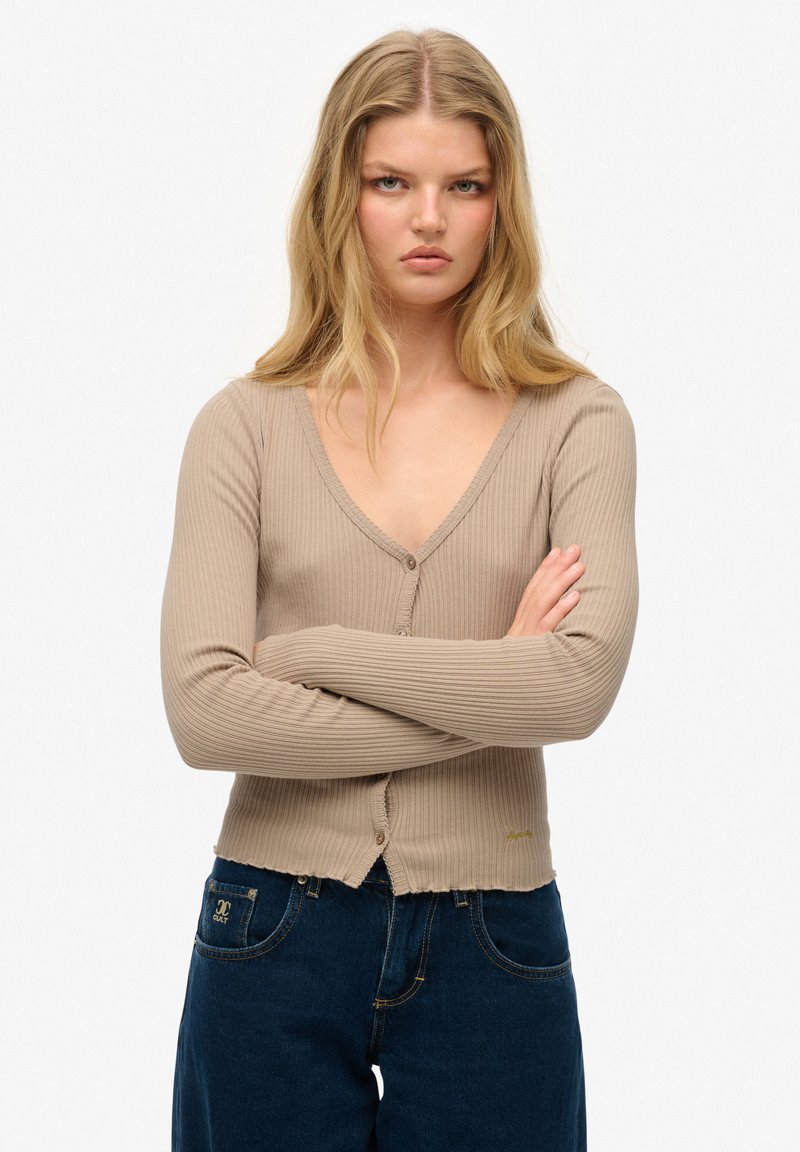 Beżowy kardigan ze strukturalnym wzorem, z głębokim dekoltem w kształcie litery V, długimi rękawami, z przodem zapinanym na guziki i postrzępionym wykończeniem, w połączeniu z ciemnoniebieskimi dżinsami.