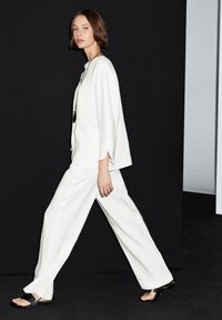 Weißes Leinenoutfit mit locker sitzendem Oberteil mit weiten Ärmeln und weitem Hosenbein. Modell trägt flache Sandalen und steht vor einem dunklen Hintergrund.