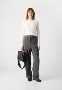 Witte kabelgebreide trui, hooggerimpelde grijze broek en een zwarte handtas met gouden accenten. Soepele textuur met een getailleerde pasvorm.