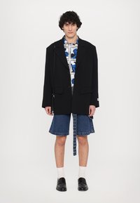 Black oversized blazer over a blue and white patterned shirt, paired with denim shorts with a snap detail, white socks, and black shoes.