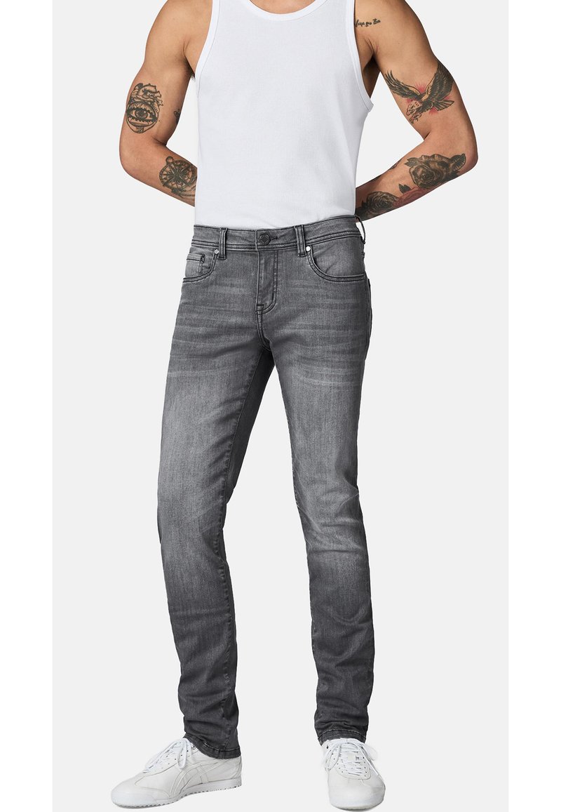 Hombre con vaqueros grises ajustados, camiseta sin mangas blanca, zapatillas blancas, con los brazos tatuados, de pie con las manos detrás de la espalda sobre un fondo blanco.