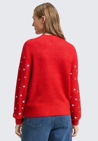 Roter Pullover aus weichem Material, mit rundem Halsausschnitt, langen Ärmeln und weißen Herzmustern an den Ärmeln. Auffällig strukturiertes Gewebe.