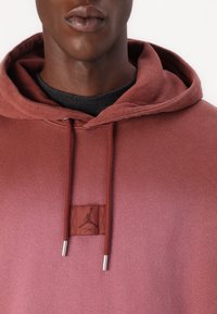 Marsala-colored hoodie made of soft fabric, featuring a metal-tipped drawstring and a logo patch near the neckline. Smooth texture, casual design.