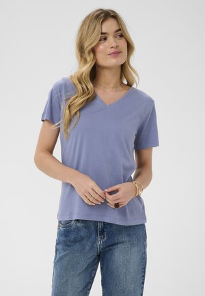 Lyshåret kvinde i lyseblå T-shirt med V-hals og blå jeans, stående med hænderne foldede, kigger let til siden mod en ensfarvet baggrund.