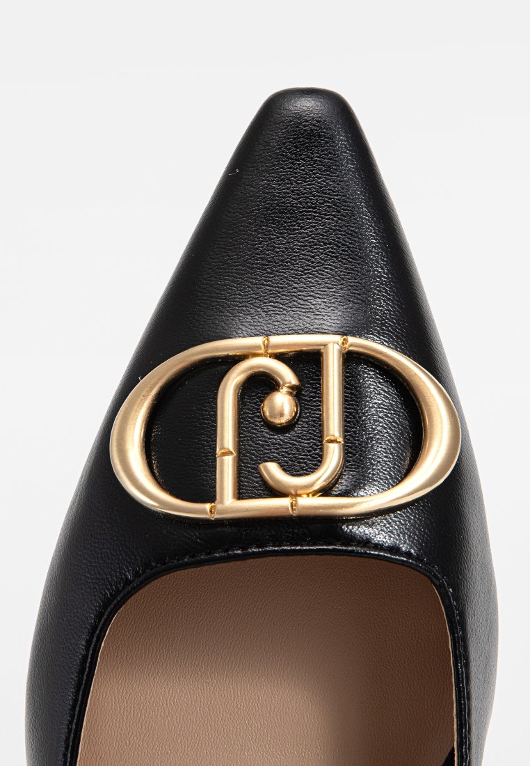 Zapato negro de piel con punta afilada que presenta un destacado acento de metal dorado con un diseño circular y un detallado intrincado. Textura suave.