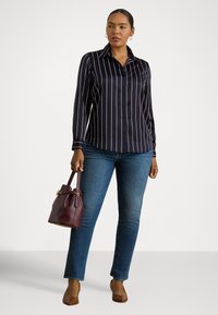 Žena v čiernej pruhovanej blúzke s dlhým rukávom, modrých džínsoch, hnedých podpätkoch, držiaca malú bordovú kabelku, stojaca pred jednoduchým pozadím.