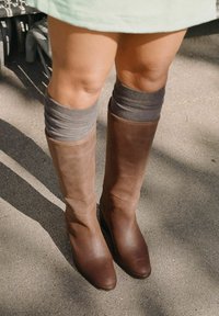 Botas de cuero marrón hasta la rodilla con un acabado liso, que presentan un panel marrón más claro en la parte superior y acentos de calcetines de rayas grises.