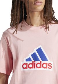 Rosa Baumwoll-T-Shirt mit einem großen weißen, blauen und roten Adidas-Logo in einem dreieckigen Design auf der Vorderseite. Kurze Ärmel und Rundhalsausschnitt.