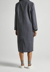 Abrigo de mezcla de lana gris con corte recto, ventilación trasera y puños abotonados, combinado con zapatillas blancas y calcetines grises acanalados.