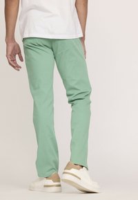 Pantalon vert clair à coupe slim et texture lisse, associé à des baskets blanches et beige. Taille et poches visibles.