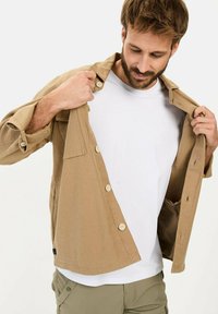 Beige katoenen shirt met knoopsluiting en borstzak, gedragen over een wit T-shirt, met opgerolde mouwen en een relaxte pasvorm.