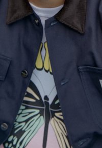 Marineblaue Cord-Jacke mit schwarzem Kragen; getragen über einem hellblauen T-Shirt mit einem großen Schmetterlings-Design in Grün, Gelb und Pink.