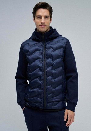 Uomo che indossa una giacca trapuntata blu navale con zip, maniche unite e cappuccio, in piedi davanti a uno sfondo grigio chiaro uniforme.