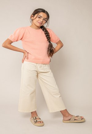 Jeune femme aux longs cheveux tressés portant un pull pêche à manches courtes, un pantalon large crème et des sandales beige, debout les mains sur les hanches.