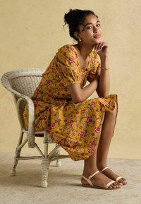 Kollane lilleline kleit puffvarrukatega, põlvepikkune, punaste ja siniste mustritega. Kanda beežide sandaalidega valgel õmblustoolil.