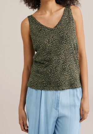 Femme portant un haut sans manches vert à imprimé léopard avec un col en V et un pantalon taille haute bleu clair, les mains détendues le long du corps.