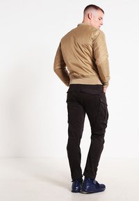 Chaqueta bomber marrón con un acabado suave, combinada con pantalones cargo negros que tienen múltiples bolsillos. Los zapatos son de color azul oscuro con un diseño deportivo.