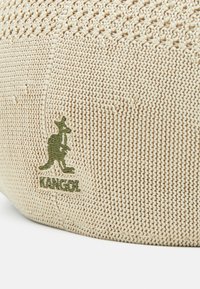 Kangol-basker i beige stickat material, med en strukturerad yta och en grön broderad logo. Rund form med platt krona.