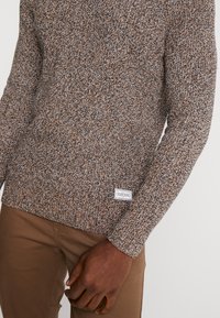 Maglione a maglia in marrone, crema e blu scuro. Presenta un motivo testurizzato con colletto e polsini a coste, e una patch con logo all'orlo.