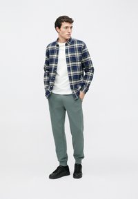 Flanellhemd in marineblauem und grün kariert, weißes T-Shirt, grüne Jogginghose und schwarze Turnschuhe, mit entspanntem Schnitt und klassischem Design.