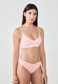 Eine Frau steht mit dem Gesicht zur Kamera und trägt einen hellrosa BH und passende Unterwäsche vor einem schlichten weißen Hintergrund.