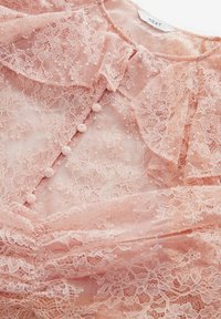Blouse en dentelle rose douce avec motifs floraux, boutons recouverts de tissu sur le devant, col à volants et détail froncé à la taille.
