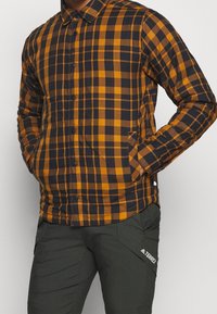 Kostkovaná košile s oranžovo-námořnickou modrou kostkovanou rysem, zapínání na knoflíky a boční kapsy. Nosená s olivově zelenými kalhotami.