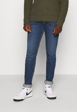 Henkilö, jolla on ylhäältä rullatut siniset farkut, oliivinvihreä neulepaita ja valkoiset Nike-tennarit mustalla swoosh-logolla, seisomassa vaalealla lattialla.