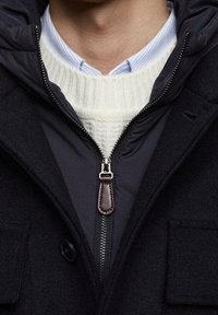 Černý vlněný kabát s kombinací materiálů, s zipem vepředu a koženým akcentem. Pod ním bílý pletený svetr z vlny, vrstvený přes světle modrou pruhovanou košili.