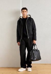 Giacca nera con cappuccio foderato in pile, abbinata a un maglione e pantaloni neri. Tiene una borsa in pelle nera e indossa scarpe da ginnastica bianche.