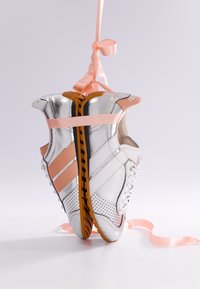 Paire de chaussures de sport argentées et blanches avec des rayures roses suspendues par des rubans roses sur un fond clair uni.