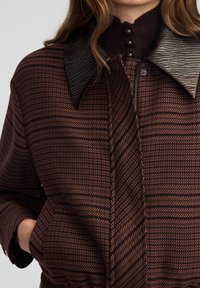 Cappotto a motivi marrone e bordeaux con un ampio colletto testurizzato, bottoni neri e un tessuto morbido. Presenta linee diagonali e orizzontali.