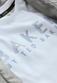 Bela bomba iz šcotne tkanine z napisom "BAKER by TED BAKER" v teksturiranem grafičnem tisku, z modrimi in črnimi črtami, leži na sivi tkanini.