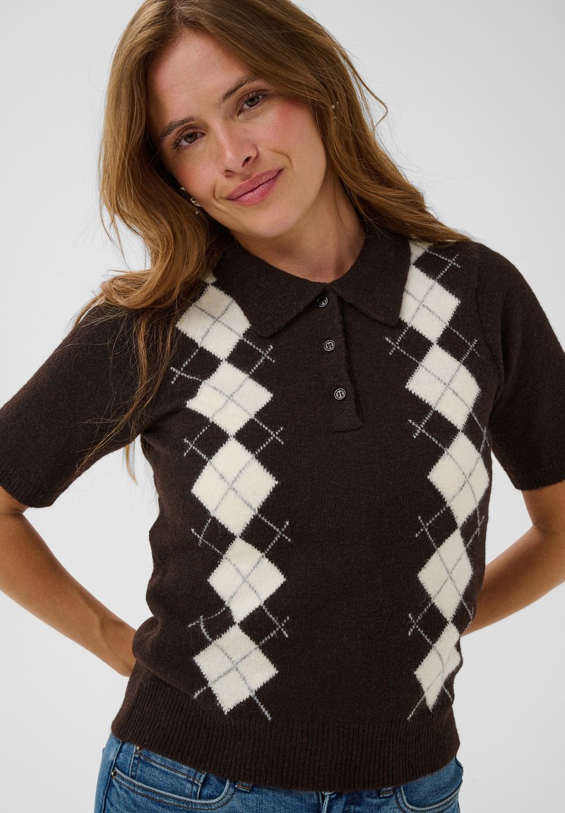 Femme aux longs cheveux châtain clair portant un pull argyle marron à manches courtes avec motif de losanges blancs, debout et souriant légèrement.