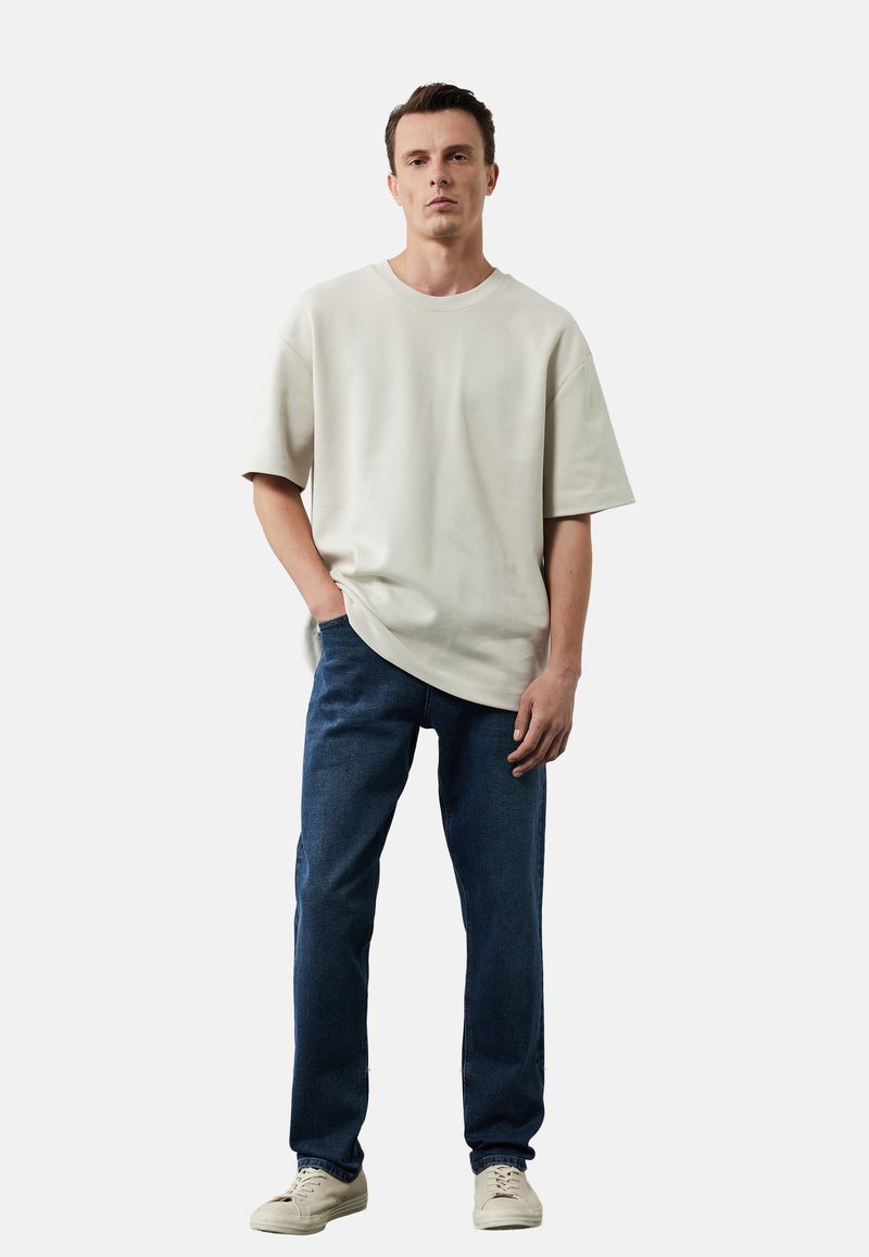 Beżowy, oversize t-shirt z krótkimi rękawami, w połączeniu z ciemnoniebieskimi dżinsami. Model stoi w luźnej pozie, ręce w kieszeniach, w jasnych sneakersach.