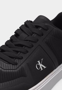 Μαύρο αθλητικό παπούτσι με υφασμάτινη υφή, μαύρα κορδόνια, λευκό λογότυπο "cK" στο πλάι και λευκή σόλα σε γκρι φόντο.