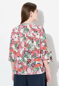 Blusa con estampado floral que presenta mangas cortas con puños atados, hecha de tejido ligero. Los colores brillantes incluyen rosa, verde, naranja y crema.