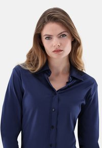 Giovane donna con occhi blu e capelli castano chiaro indossa una camicia azzurra con bottoni su uno sfondo chiaro e semplice.
