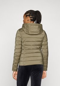 Doudoune vert olive avec capuche, présentant des sections matelassées horizontales et une texture lisse. Portée avec un pantalon en velours noir.