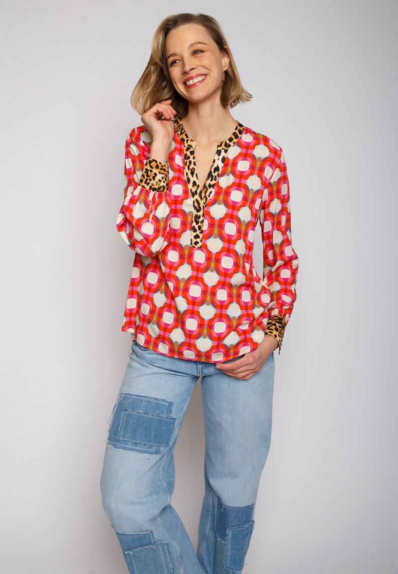 Rote, gemusterte Bluse mit kreisförmigen Designs und Leopardenmusterakzenten am Kragen und an den Manschetten. Kombiniert mit hellblauen, gefleckten Jeans.