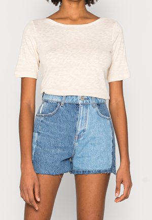 Top corto beige chiaro a maniche corte abbinato a un paio di pantaloni denim bicolore—blu scuro e blu chiaro—con vestibilità regolare, cuciture in vista e orli sfrangiati.
