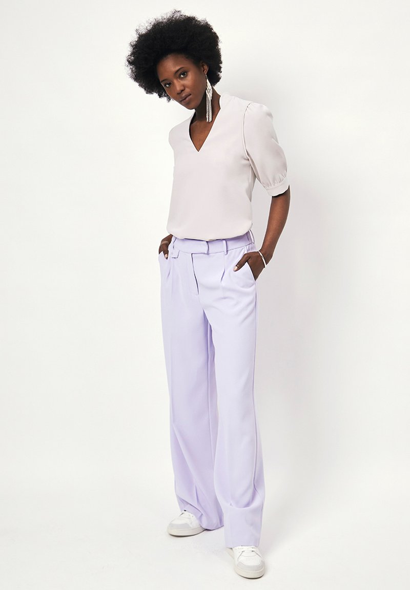 Lichtgrijze blouse met korte mouwen en een V-hals; gecombineerd met hooglopende, wijde lavendelkleurige broek; witte sneakers; stijlvolle hangoorbellen.