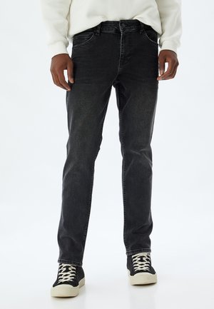 Jeans de mezclilla negra con corte ajustado, cinco bolsillos y una sutil textura desgastada. Combinados con zapatillas negras con cordones y suela blanca.